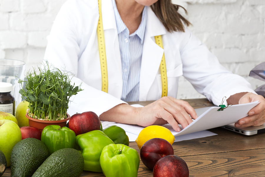 Cropped image of unrecognizable mature female dietician in white medical gown holding pen and papers, studying medical records of client, sitting at desk with veggies. Selective focus on woman's hands